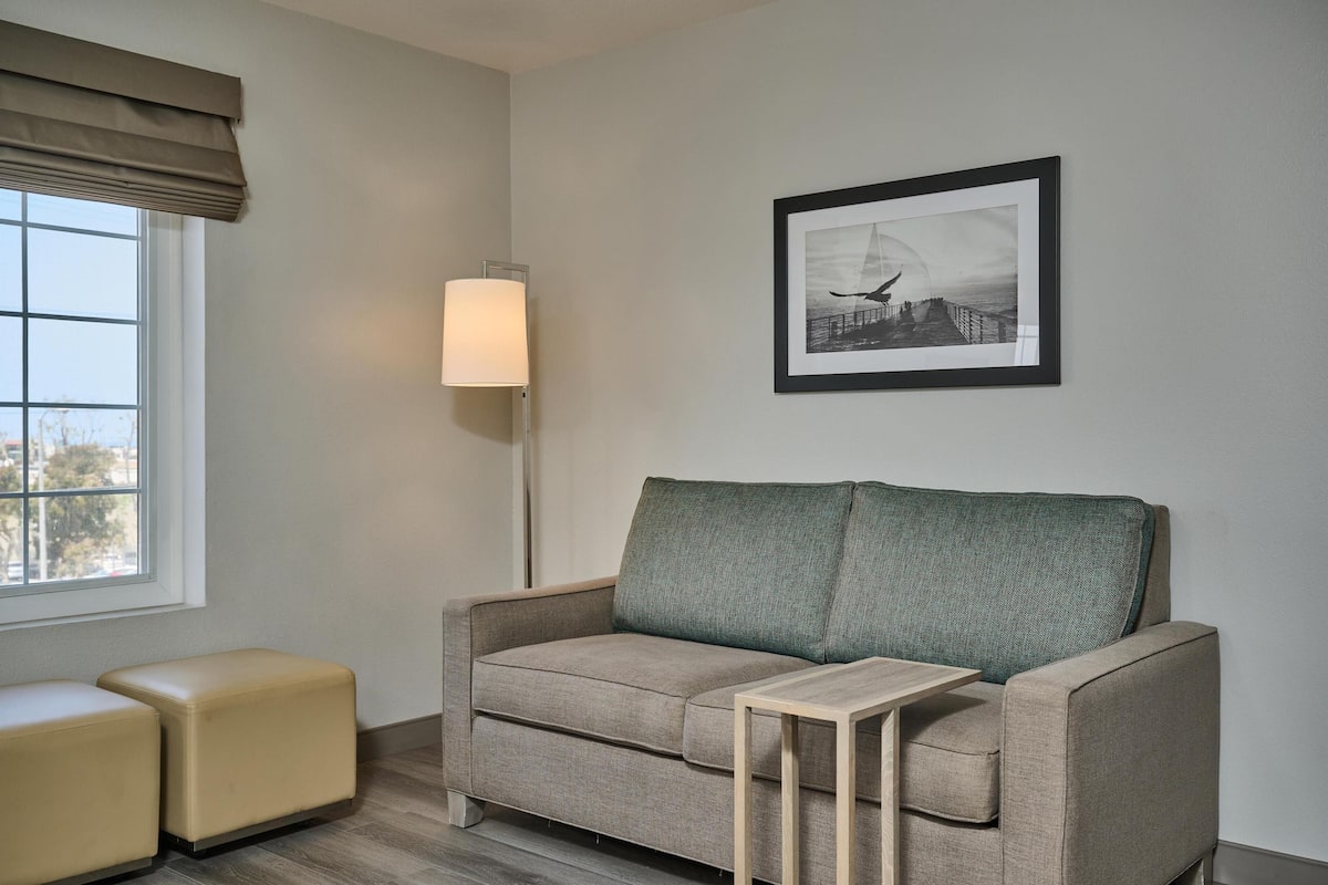 A neutral-colored sofa with a matching ottoman and a small side table, positioned in a room with a window and a framed picture on the wall.