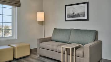 A neutral-colored sofa with a matching ottoman and a small side table, positioned in a room with a window and a framed picture on the wall.