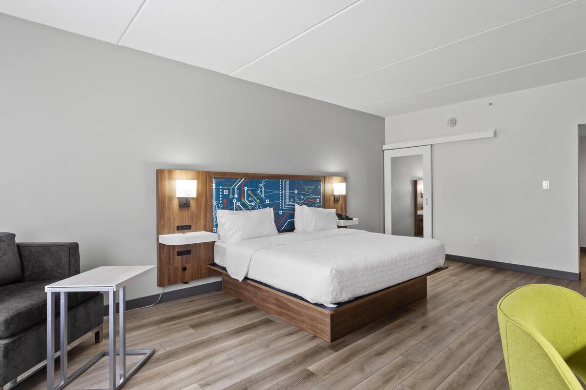 A modern hotel room with a wooden bed, bedside tables, a grey sofa, and a bright green chair.