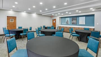 A conference room with round tables and blue chairs, equipped with ceiling lights and a wall-mounted display.