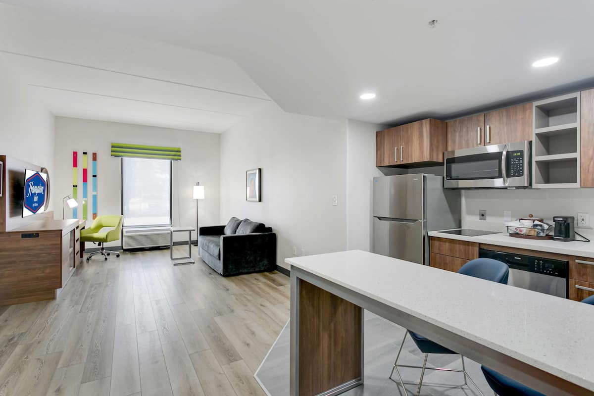 A modern kitchen with a microwave, refrigerator, and dishwasher. There is a dining area with a long table and chairs. A sofa and a chair are visible in the background.
