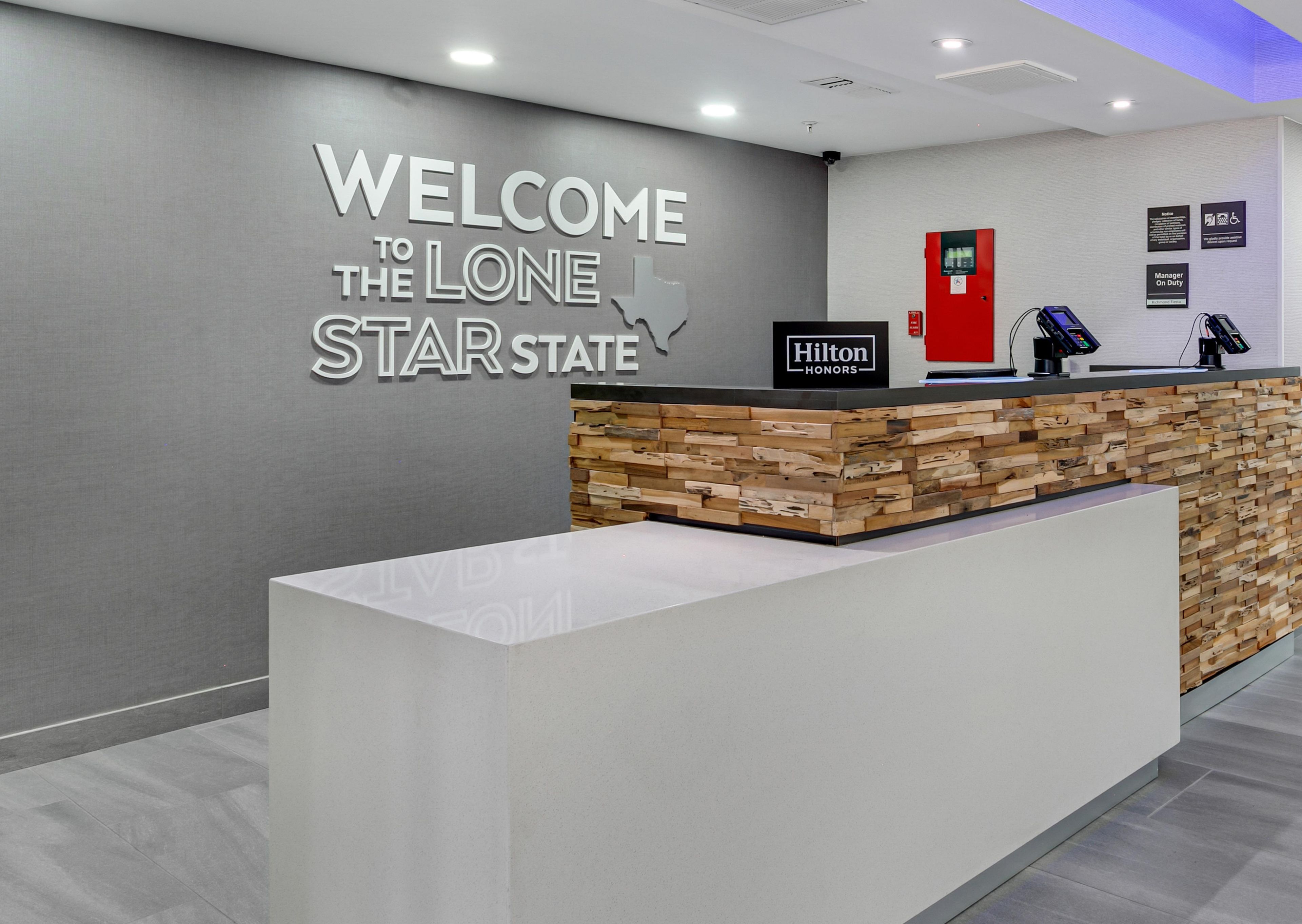Una recepción de hotel con un cartel que dice 'WELCOME TO THE LONE STAR STATE', una insignia de 'Hilton Honors' y un quiosco digital.