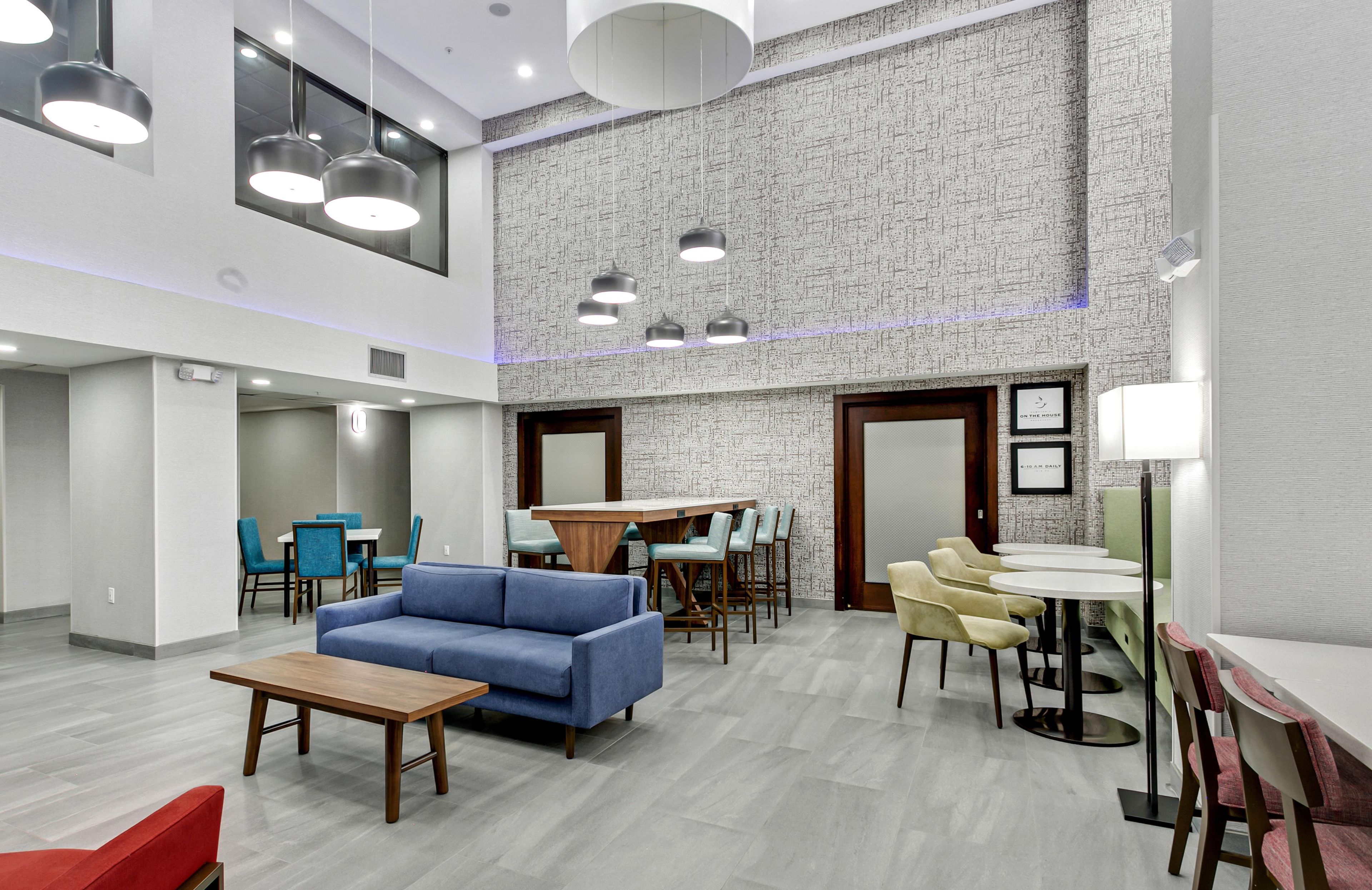 Una moderna hall d'albergo con soffitto alto, parete decorata e diverse opzioni di seduta, tra cui un divano blu, tavoli da pranzo e poltrone.