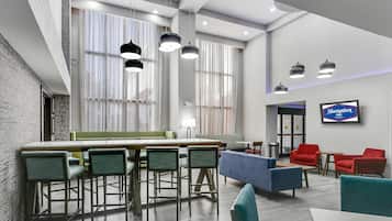A modern hotel lobby with a bar area, seating, and a television screen displaying the hotel name.