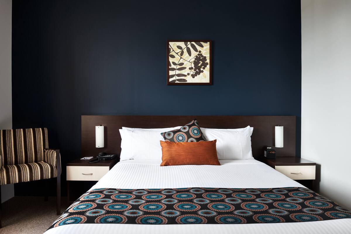 A hotel room with a bed, a nightstand, a chair, and a wall-mounted artwork.