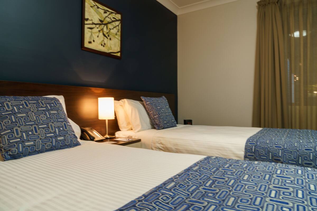 A hotel room with two beds, a nightstand, a lamp, and a painting on the wall.