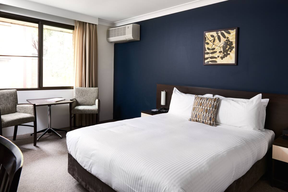 A hotel room with a large bed, a desk, two chairs, and a window with curtains.
