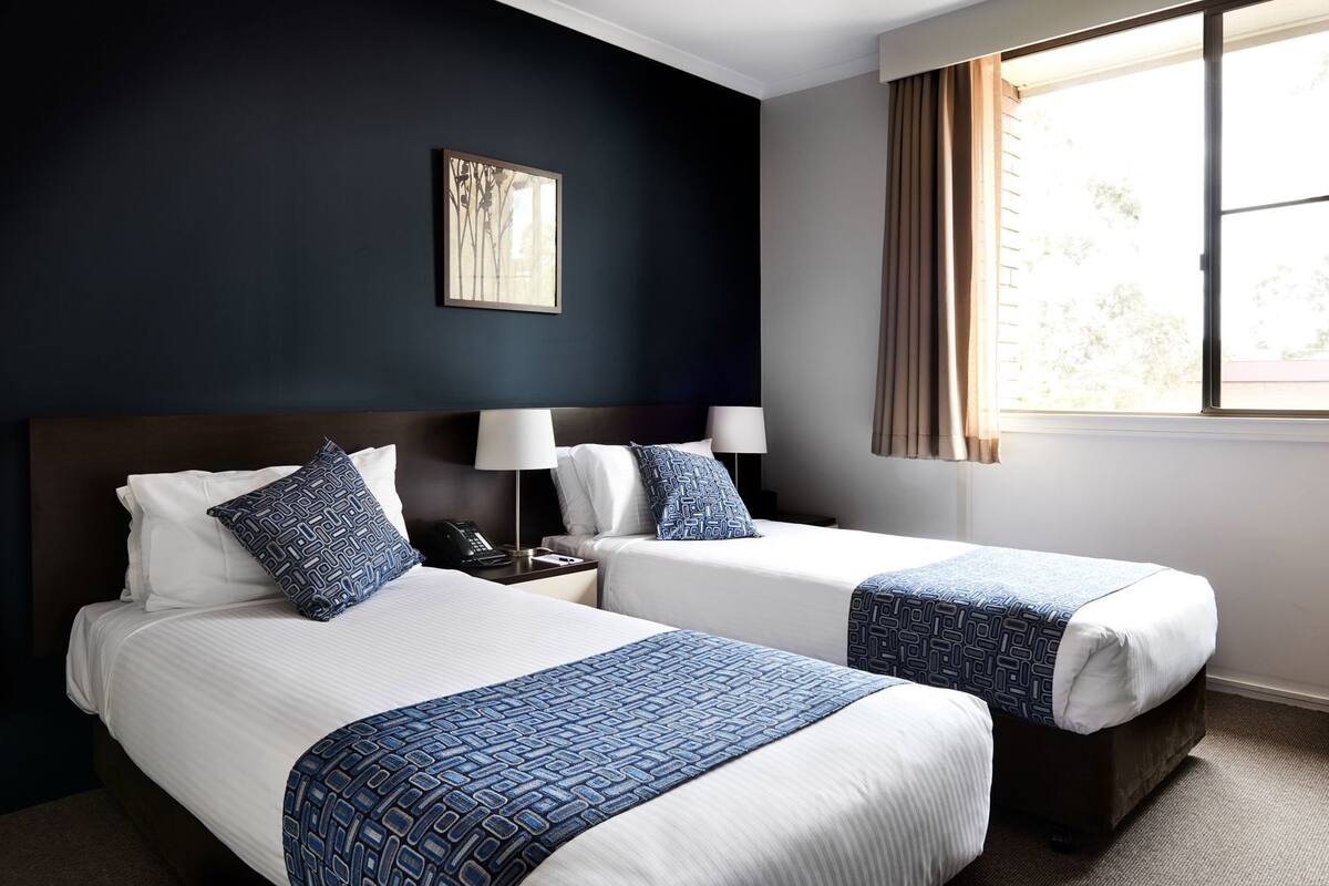 A hotel room with two beds, a nightstand with a phone, a lamp, and a window with curtains.