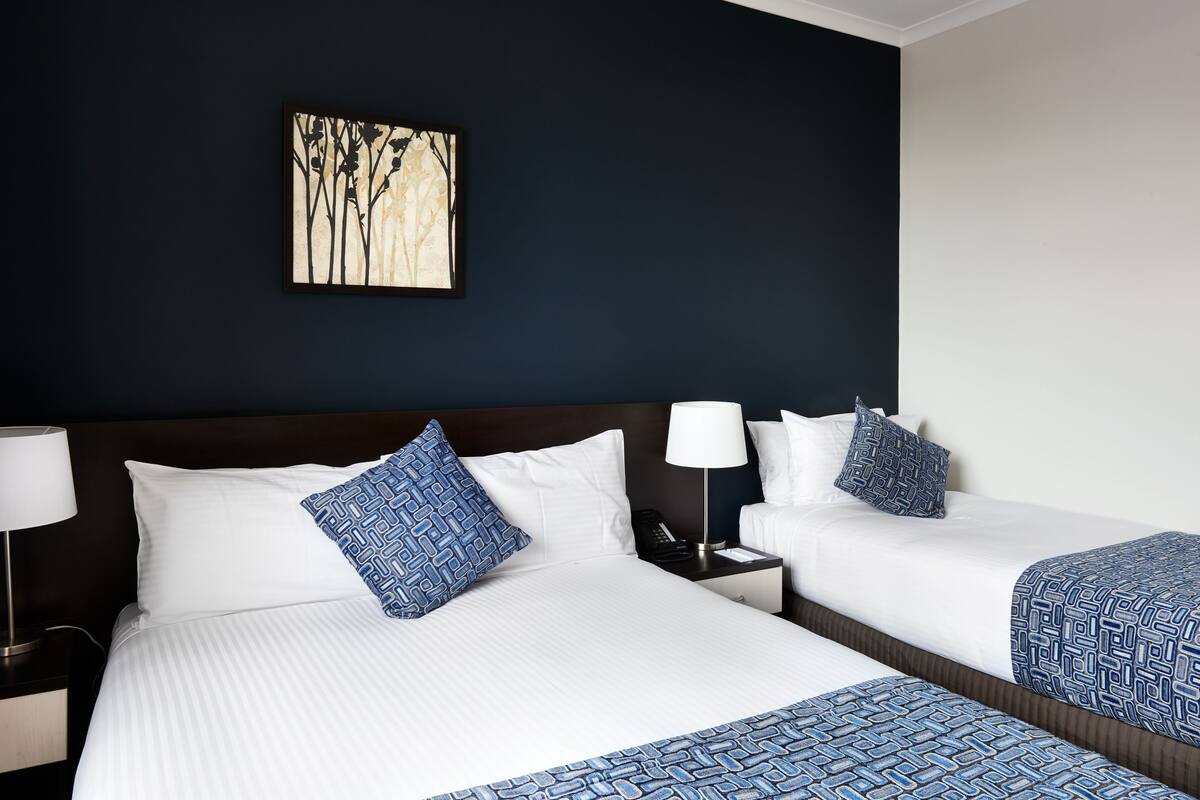 A hotel room with two beds, a nightstand with a lamp, a phone, and a wall-mounted artwork.