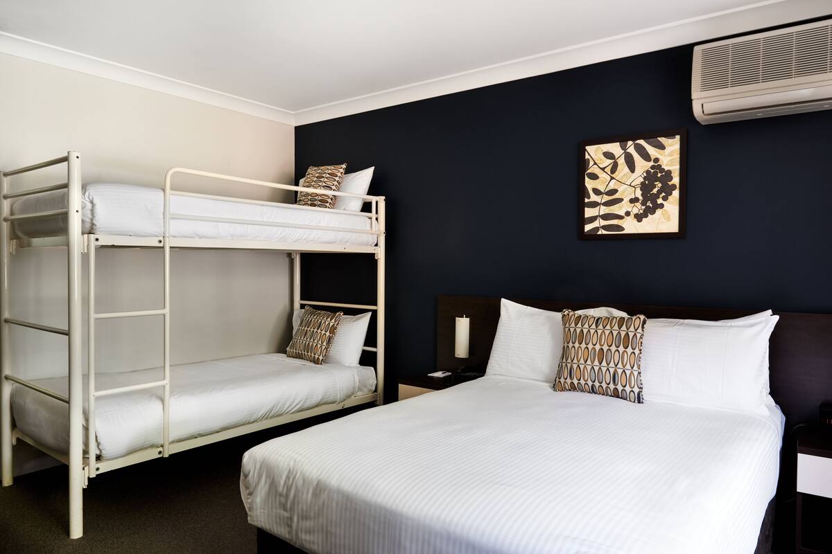 A room with a bunk bed, a single bed, a nightstand with a lamp, and a wall-mounted air conditioner.
