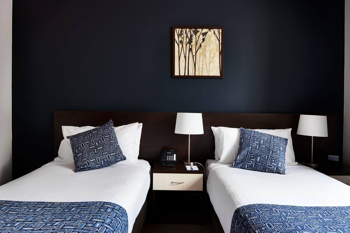 A hotel room with two beds, each with a patterned bedspread, two bedside lamps, a nightstand with a phone, and a wall-mounted artwork.