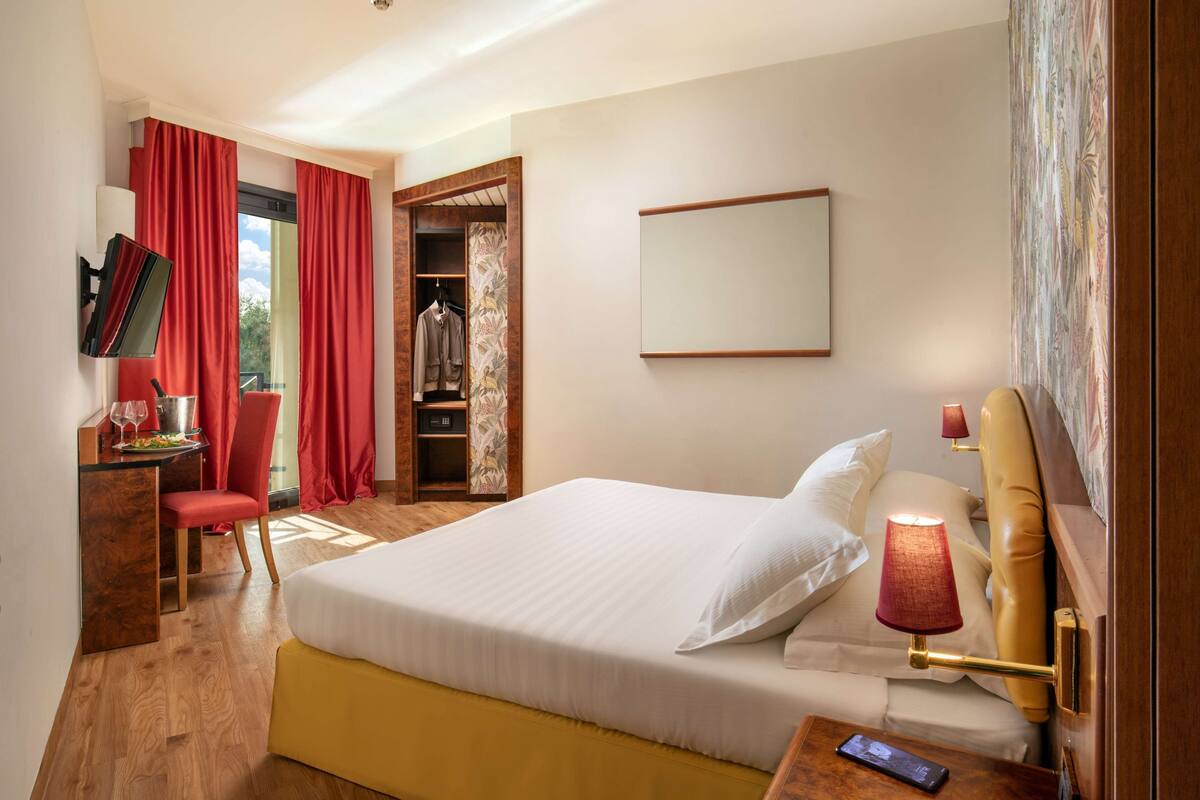 A hotel room with a bed, bedside table, lamp, and a desk with a chair and a mirror.