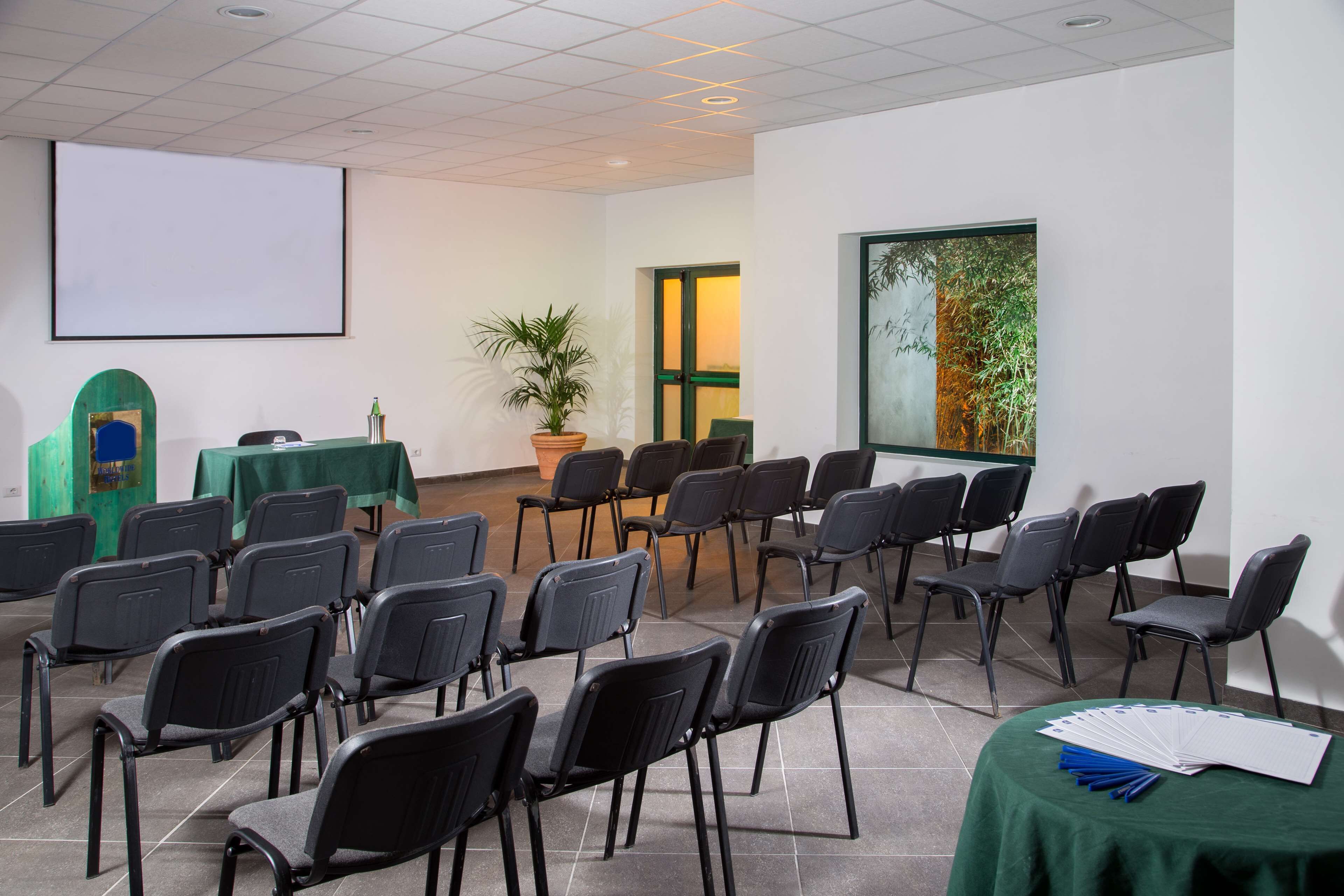 A conference room with rows of chairs, a projector screen, a table with a green tablecloth, a potted plant, and a window with a view of trees.