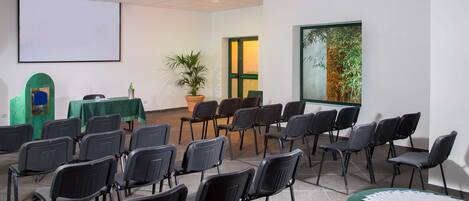 A conference room with rows of chairs, a projector screen, a table with a green tablecloth, a potted plant, and a window with a view of trees.