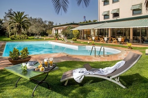 Un área junto a la piscina con una tumbona, una mesa con bebidas y un edificio con terraza.