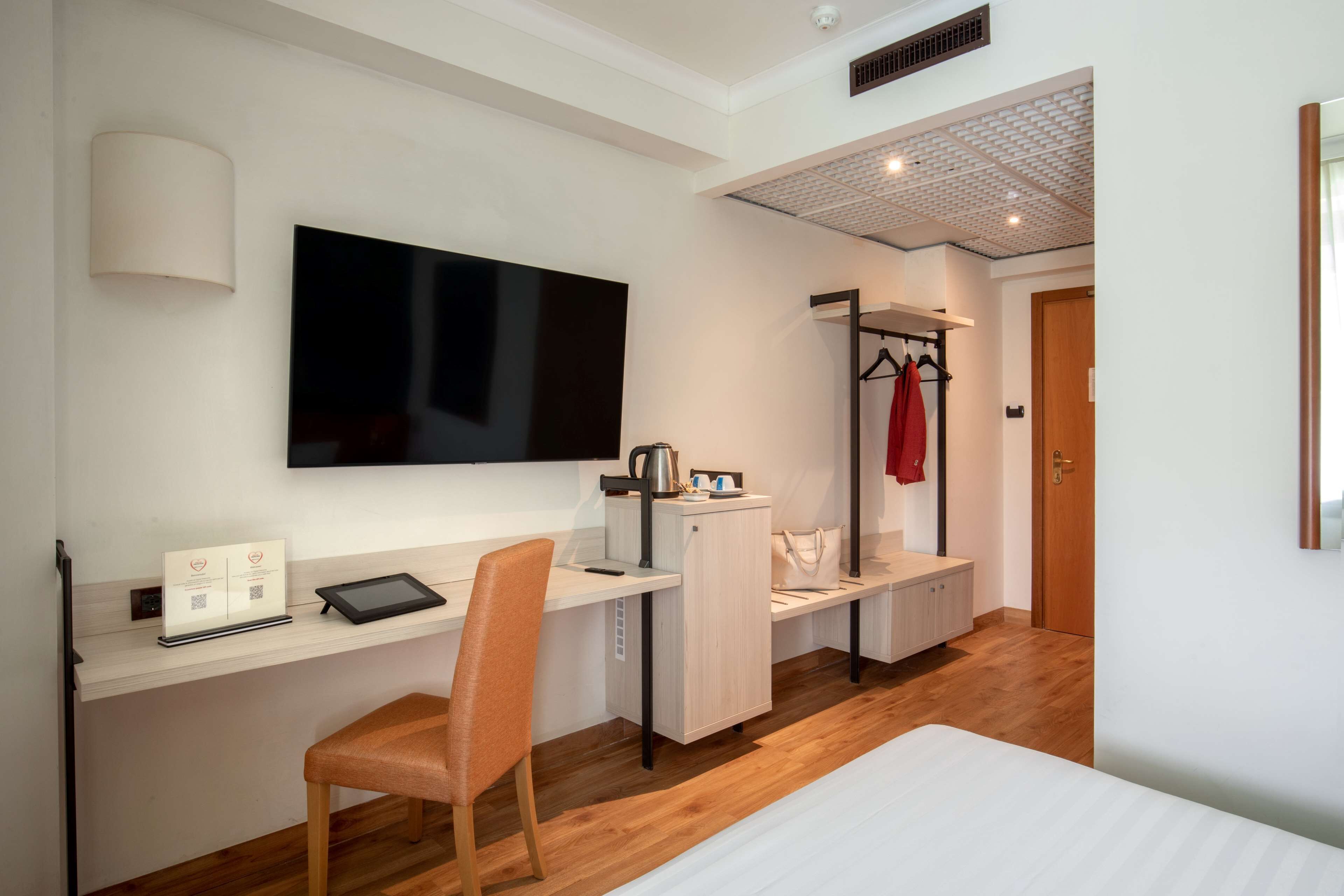 Habitación de hotel con escritorio, silla, cama, televisor, perchero y puerta.