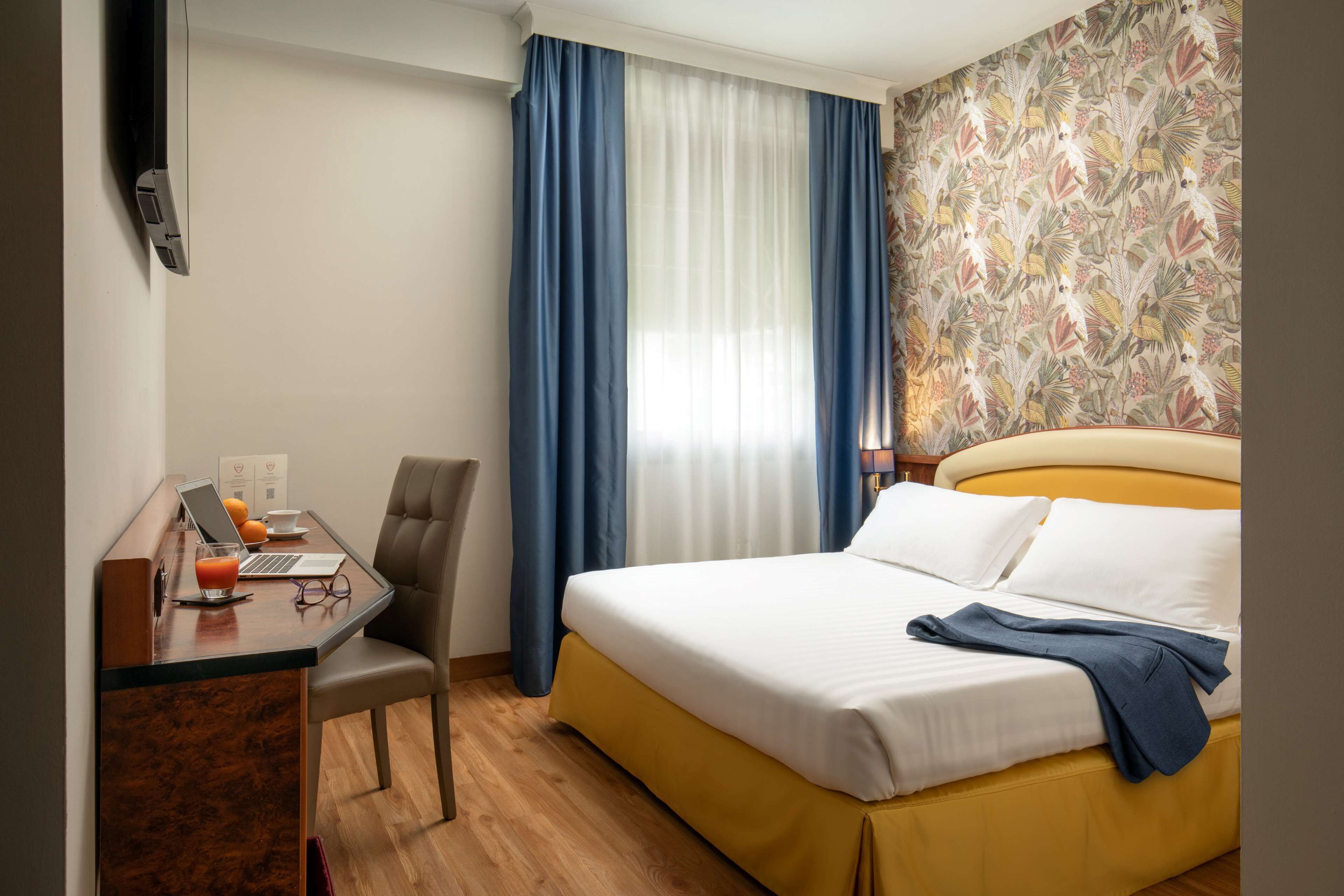 Quarto de hotel com cama, secretária com um laptop, cadeira e janela com cortinas.