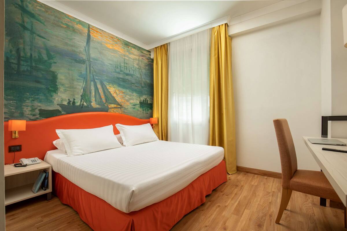 A hotel room with a bed, a desk, a chair, a nightstand, and a painting on the wall.
