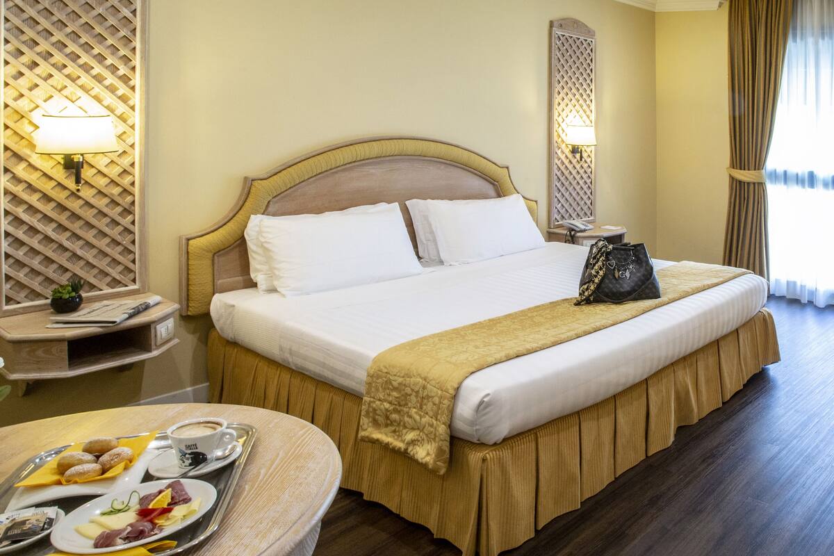 A hotel room with a large bed, a desk, a chair, and a dining area with breakfast.