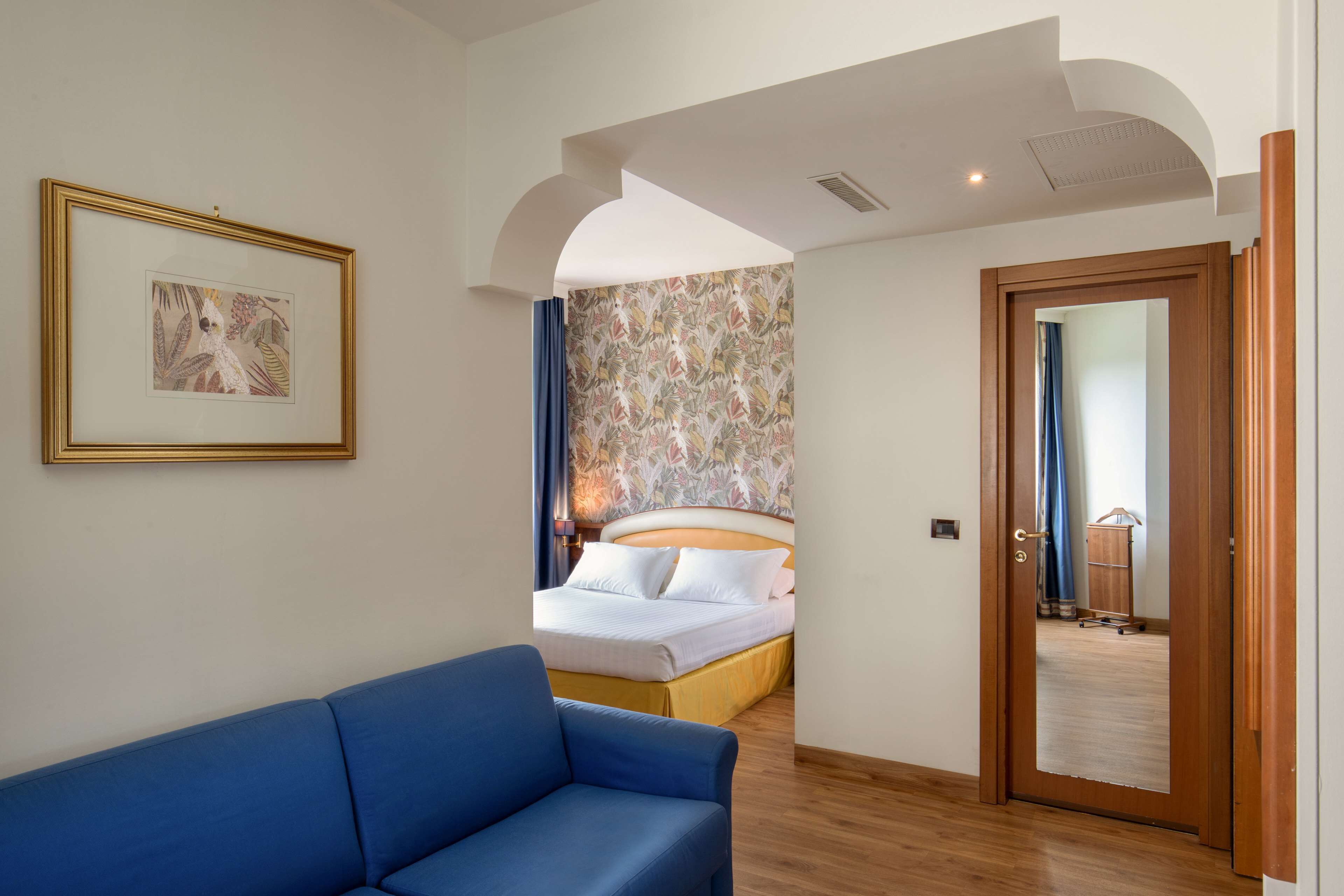 A hotel room with a bed, a blue sofa, a framed picture on the wall, and a door leading to another room.