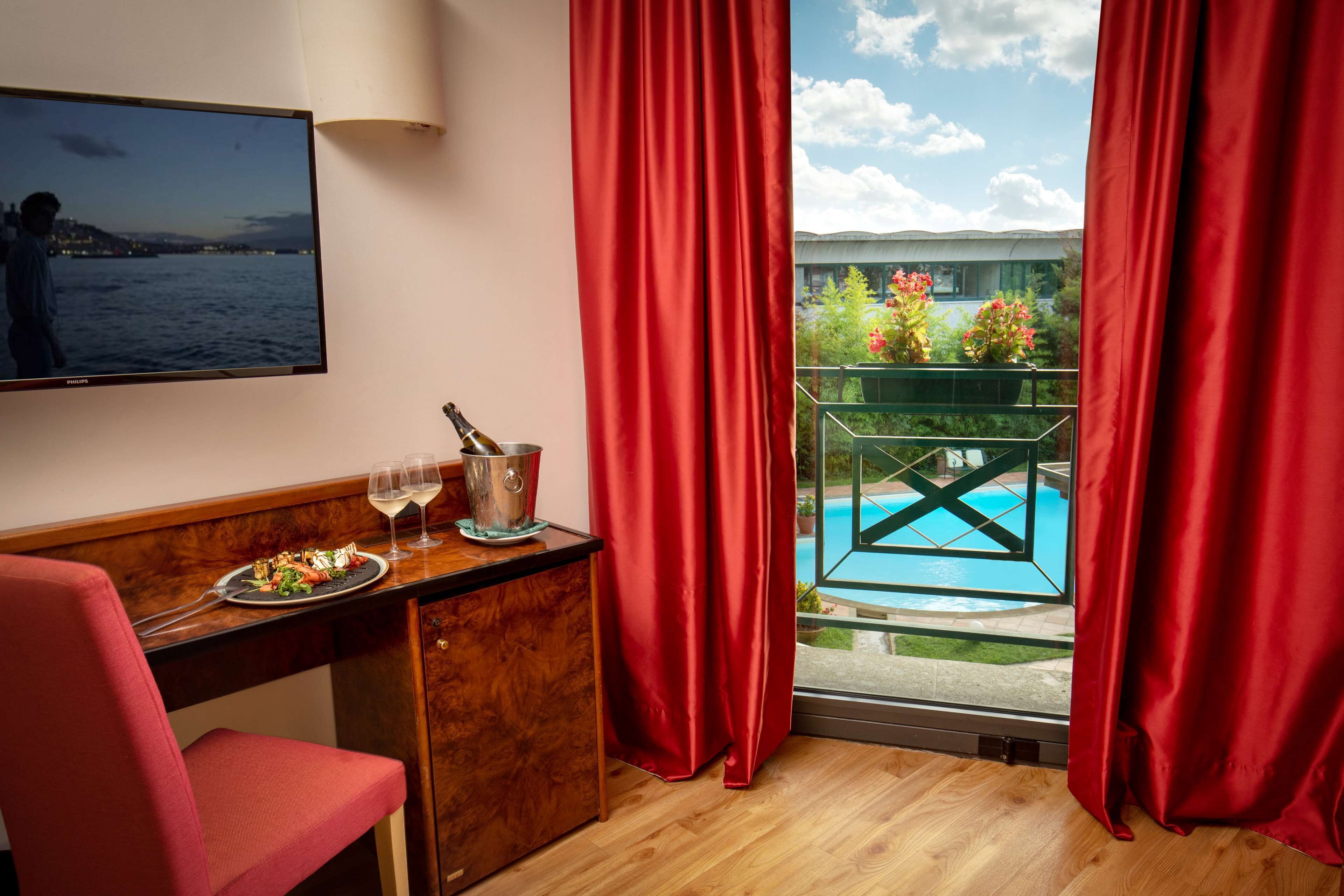 A room with a wooden desk, a red chair, a television, a bottle of champagne, and a glass of wine. There is a window with red curtains and a view of a pool and greenery.