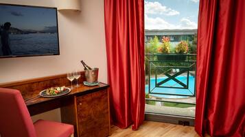 A room with a wooden desk, a red chair, a television, a bottle of champagne, and a glass of wine. There is a window with red curtains and a view of a pool and greenery.