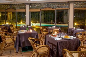 Restaurante com mesas preparadas para refeições, piscina ao fundo e grandes janelas.