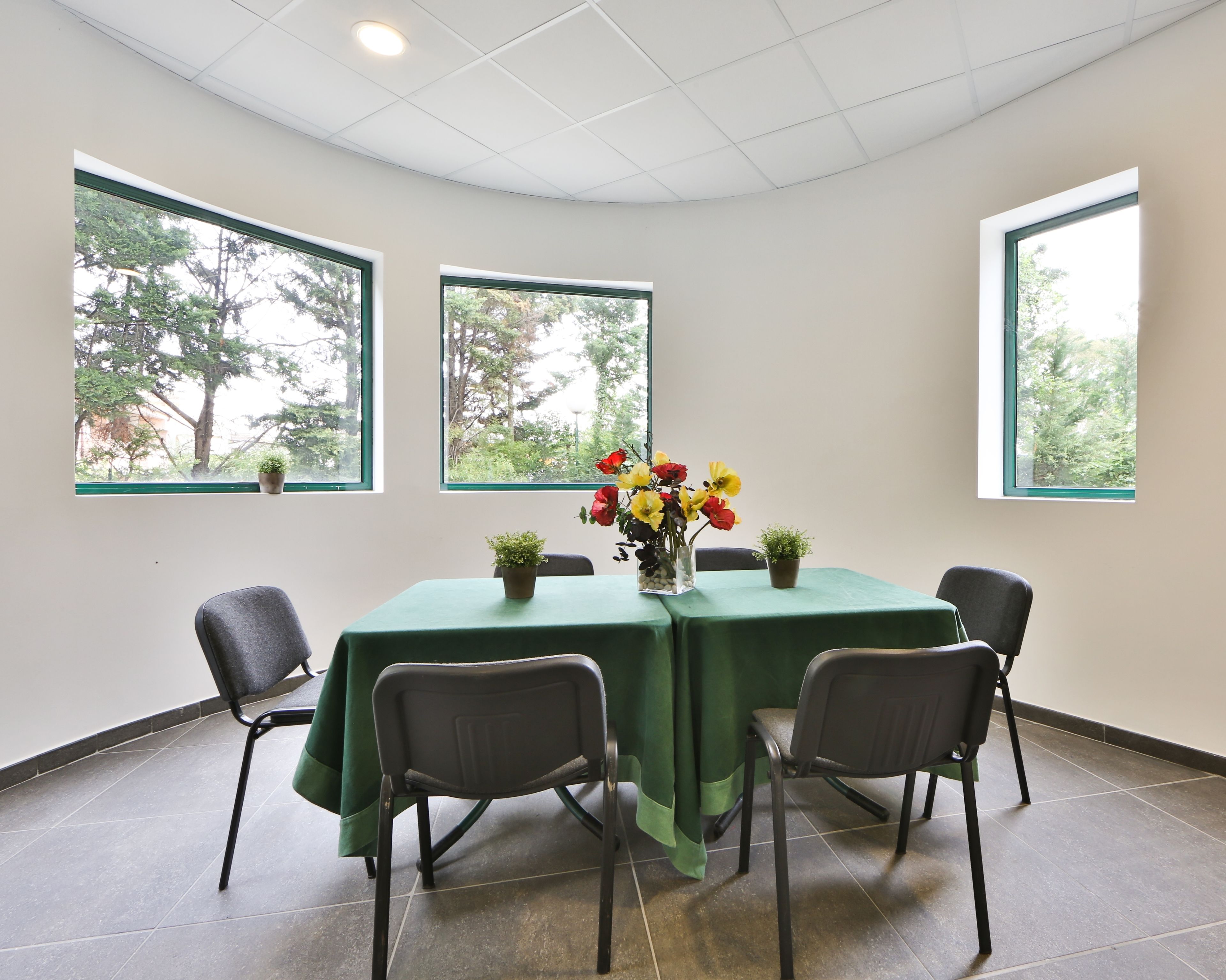 Un salón de conferencias con una mesa verde, cuatro sillas, un jarrón de flores y tres ventanas con vistas a árboles.