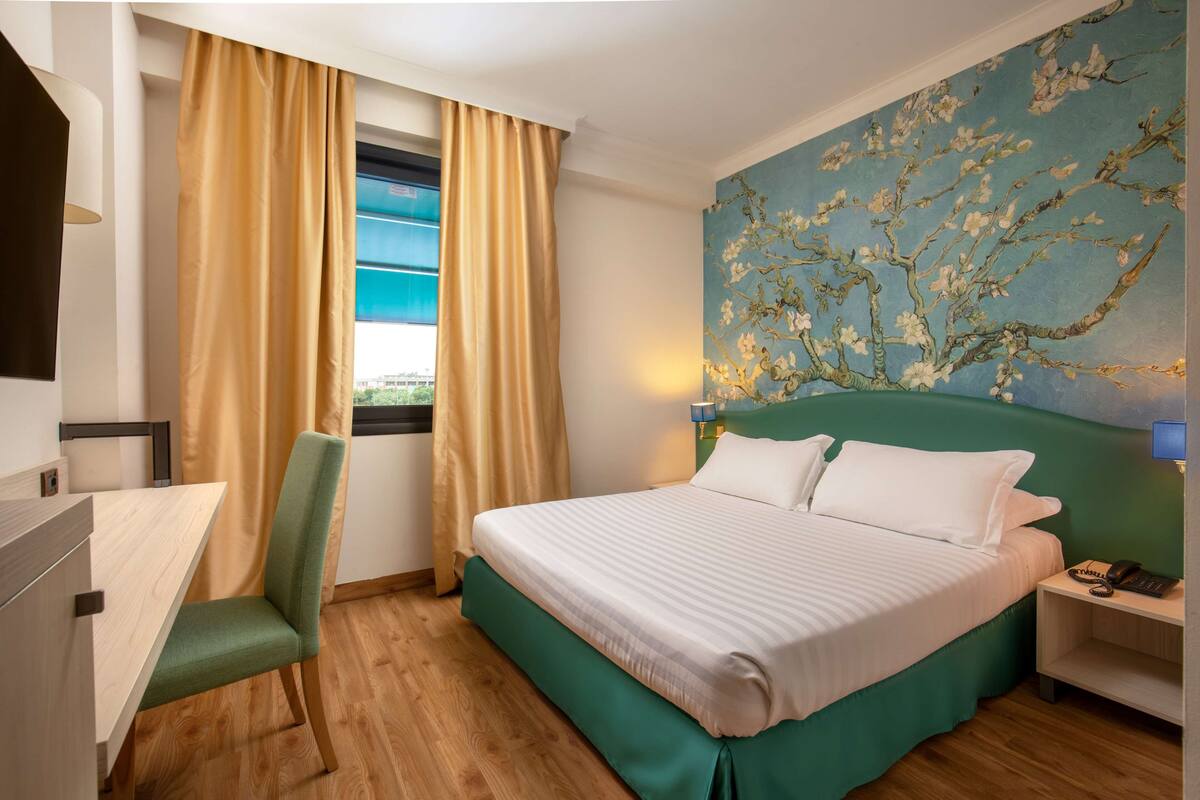 A hotel room with a bed, a desk, a chair, a nightstand, and a painting on the wall.
