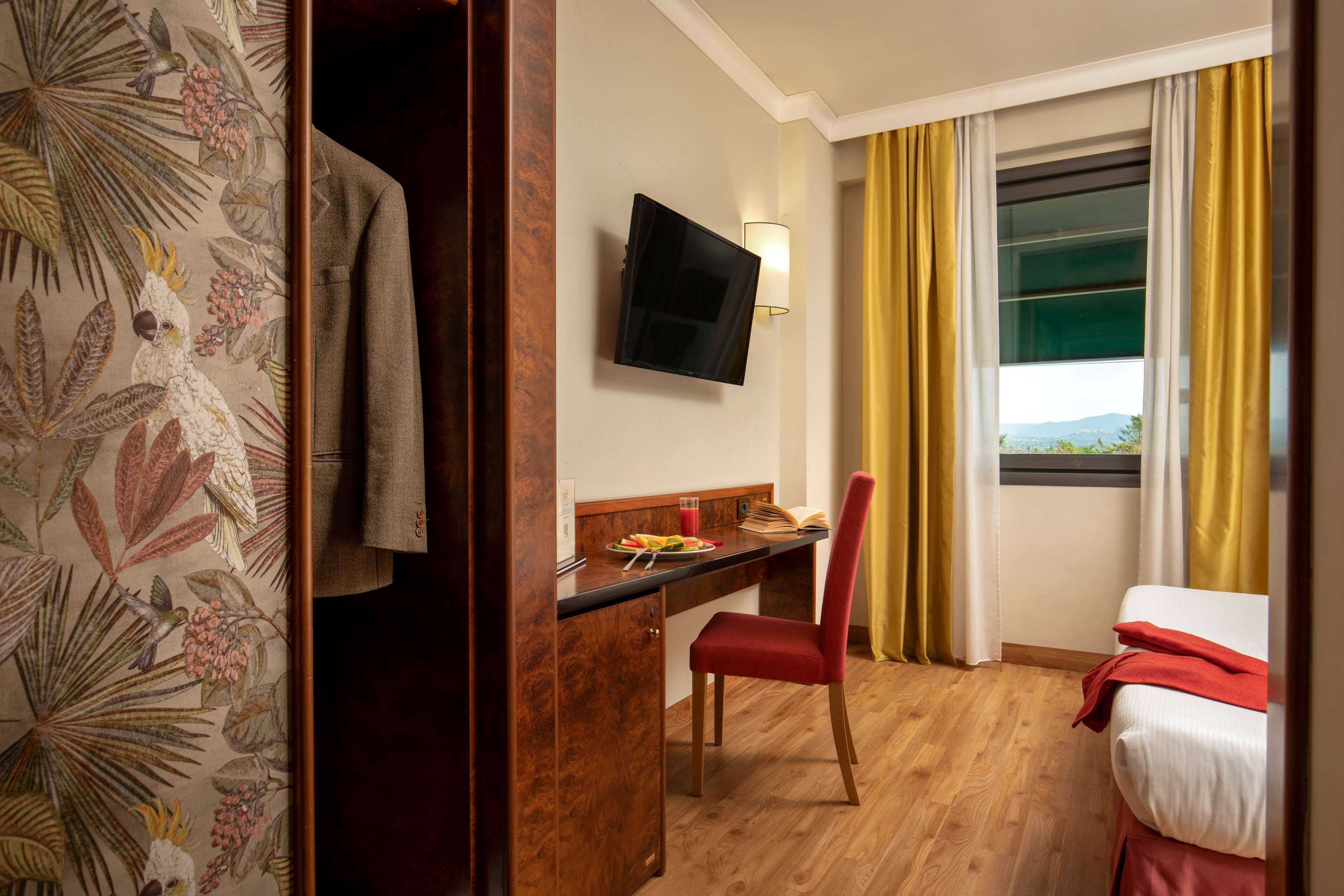 Camera d'albergo con una scrivania in legno, una sedia rossa, un letto con coperta rossa, una TV a parete e una finestra con tende gialle.