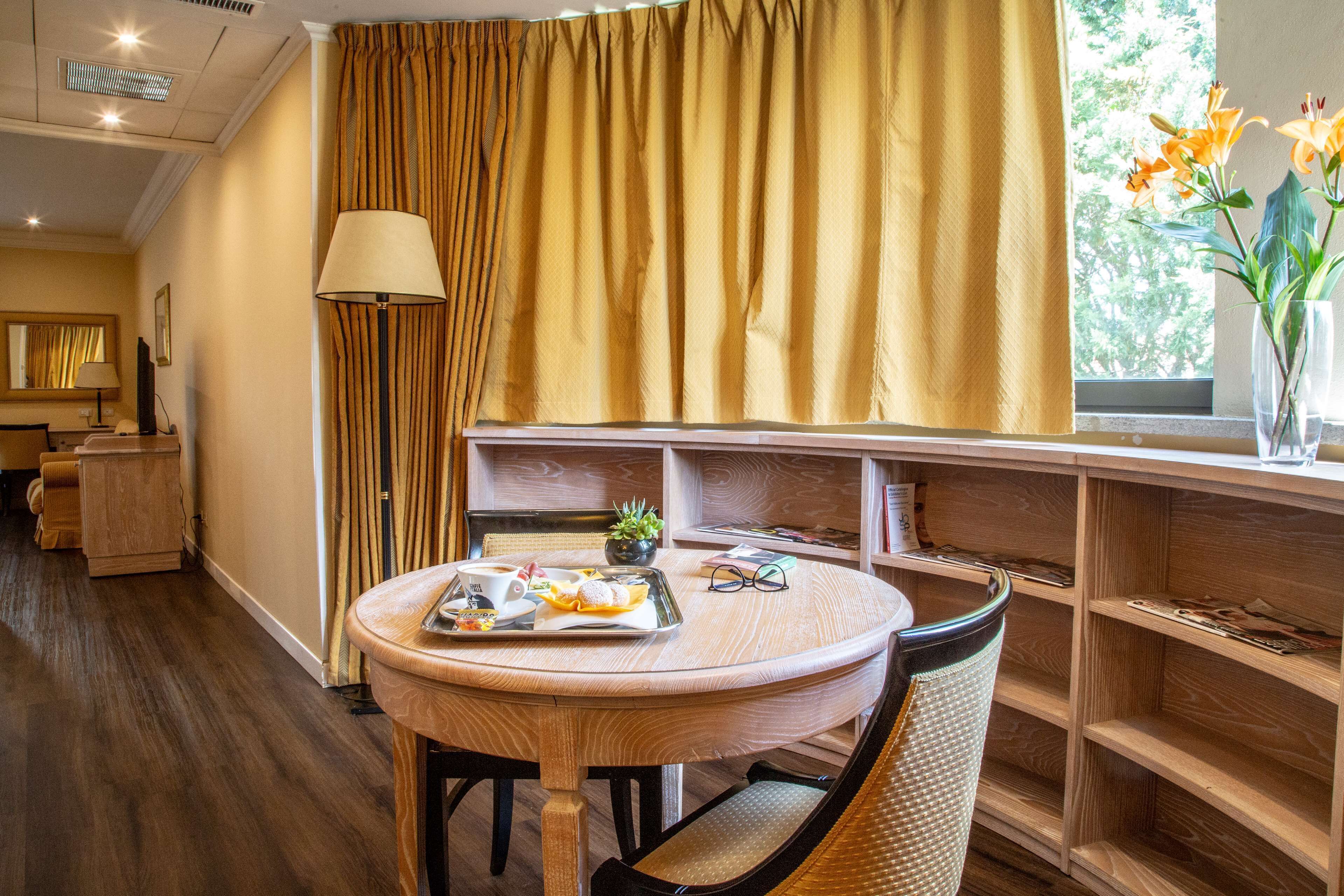 A dining area with a round table, chairs, a tray with food, a lamp, and a window with curtains.