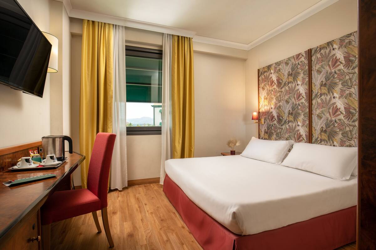 A hotel room with a bed, a desk with a chair, a television, a window with curtains, and a decorative wall panel.
