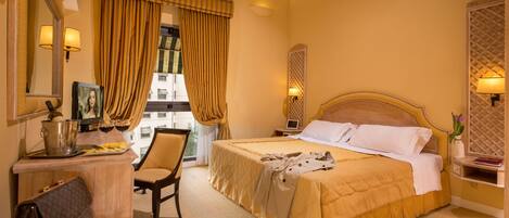 A hotel room with a bed, a desk with a laptop, a chair, a bedside table with a lamp, and a view of the outside.