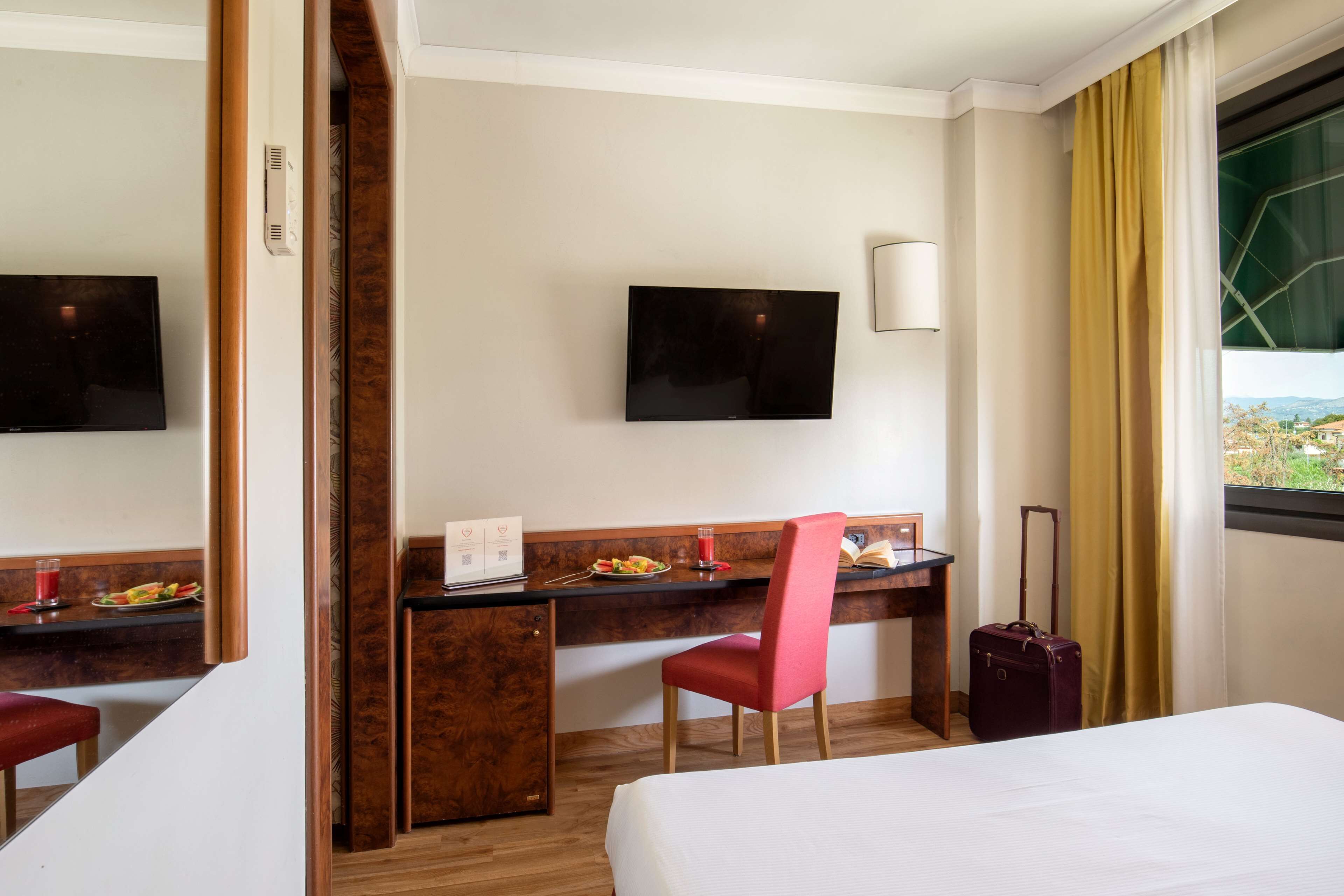 A hotel room with a bed, a desk with a chair, a television, and a window with curtains.