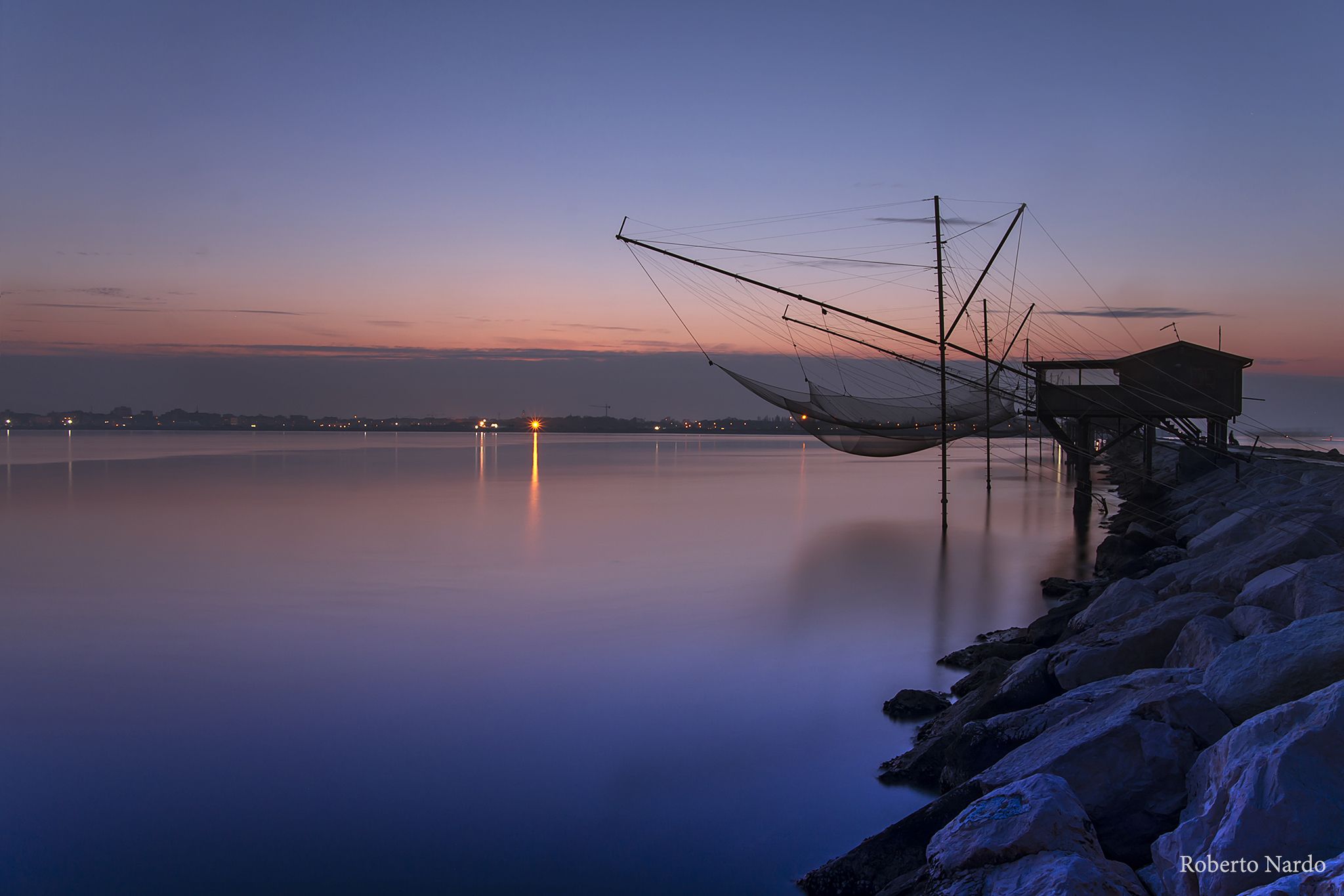 A tranquil waterfront scene with a fishing net structure and a small wooden building, likely a dock or shed, by the water's edge.