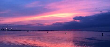 A calm body of water reflecting a sunset sky with a few structures in the distance.