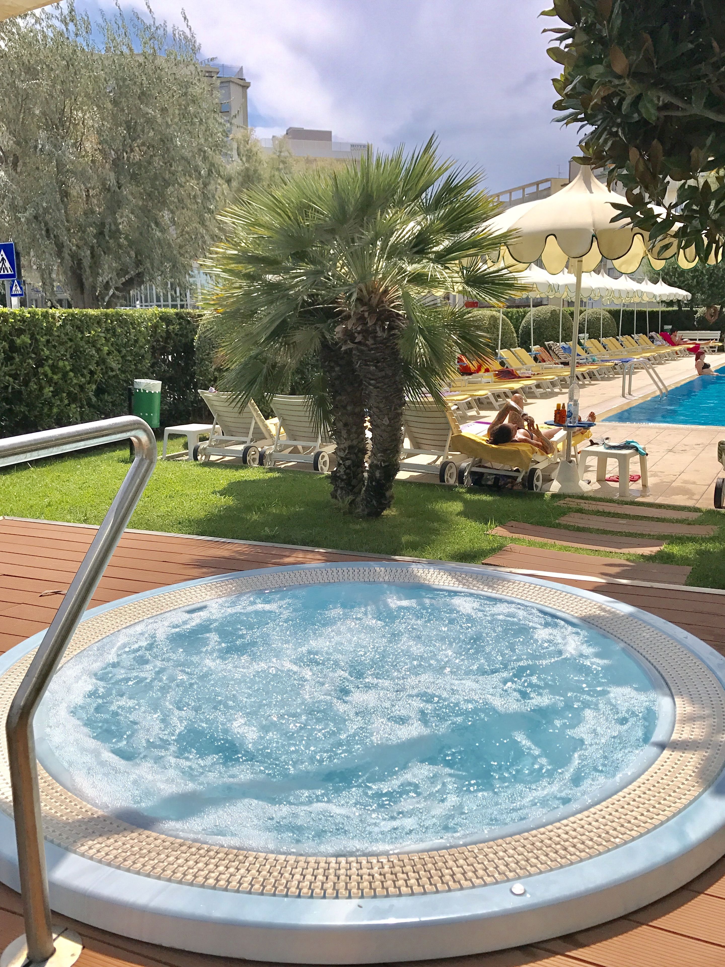 Ein Whirlpool, umgeben von Liegestühlen und Palmen, mit einem Pool und Sonnenschirmen im Hintergrund.