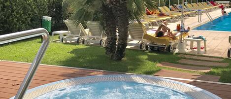 Ein Whirlpool, umgeben von Liegestühlen und Palmen, mit einem Pool und Sonnenschirmen im Hintergrund.