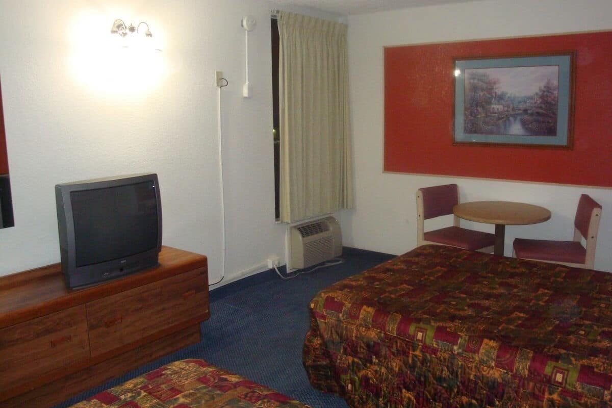 A hotel room with a bed, a TV on a wooden dresser, a small table with chairs, and a painting on the wall.