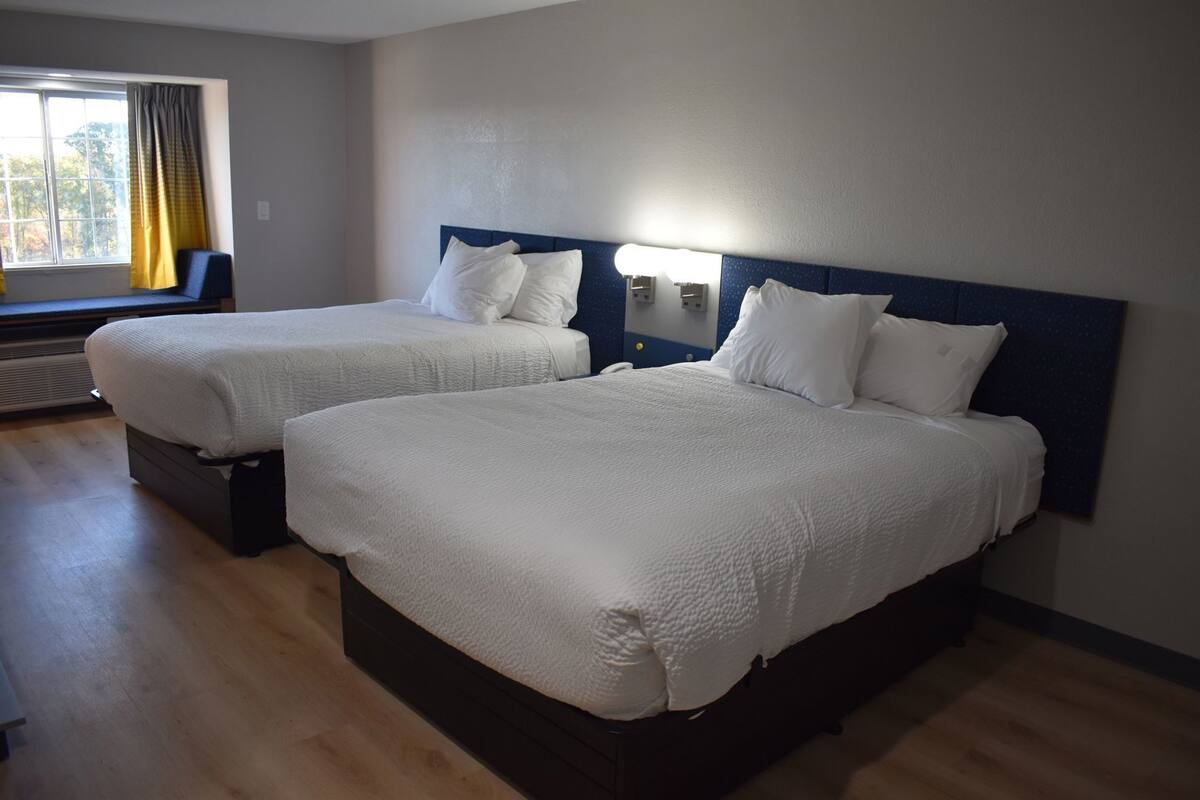 Deux lits avec du linge de lit blanc, une fenêtre avec des rideaux et un luminaire fixé au mur.