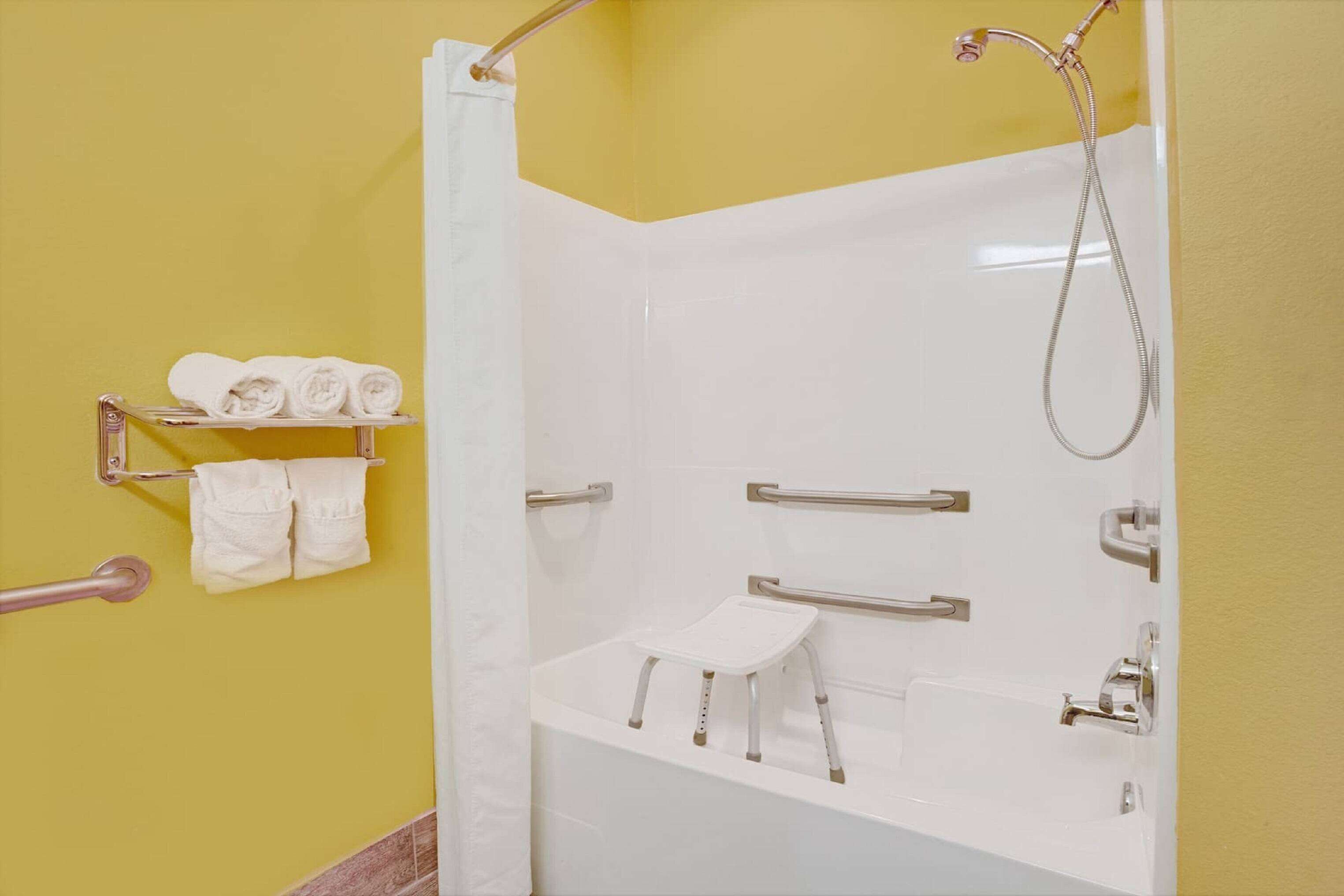 A bathroom with a white bathtub, a shower, and a small white stool. There are towels on a rack and a grab bar on the wall.