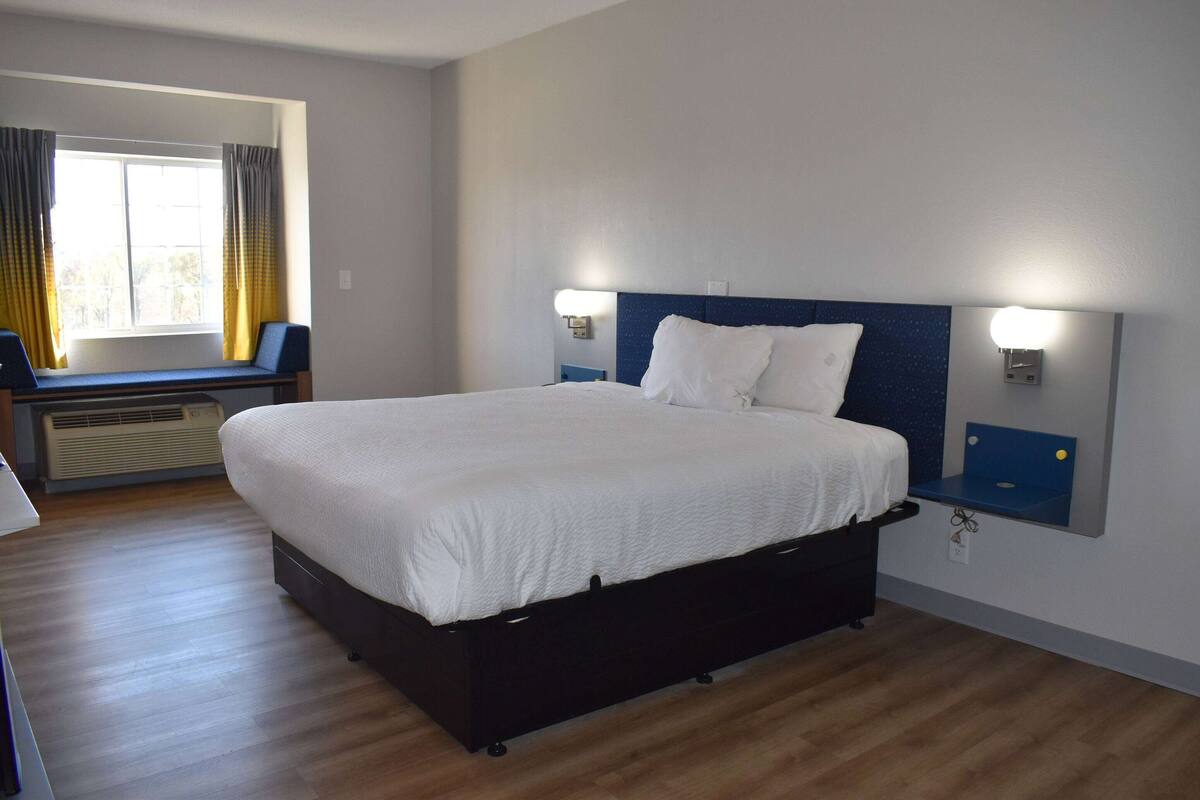 Une chambre d’hôtel avec un grand lit, une fenêtre avec des rideaux, une table de chevet avec une lampe et un luminaire fixé au mur.