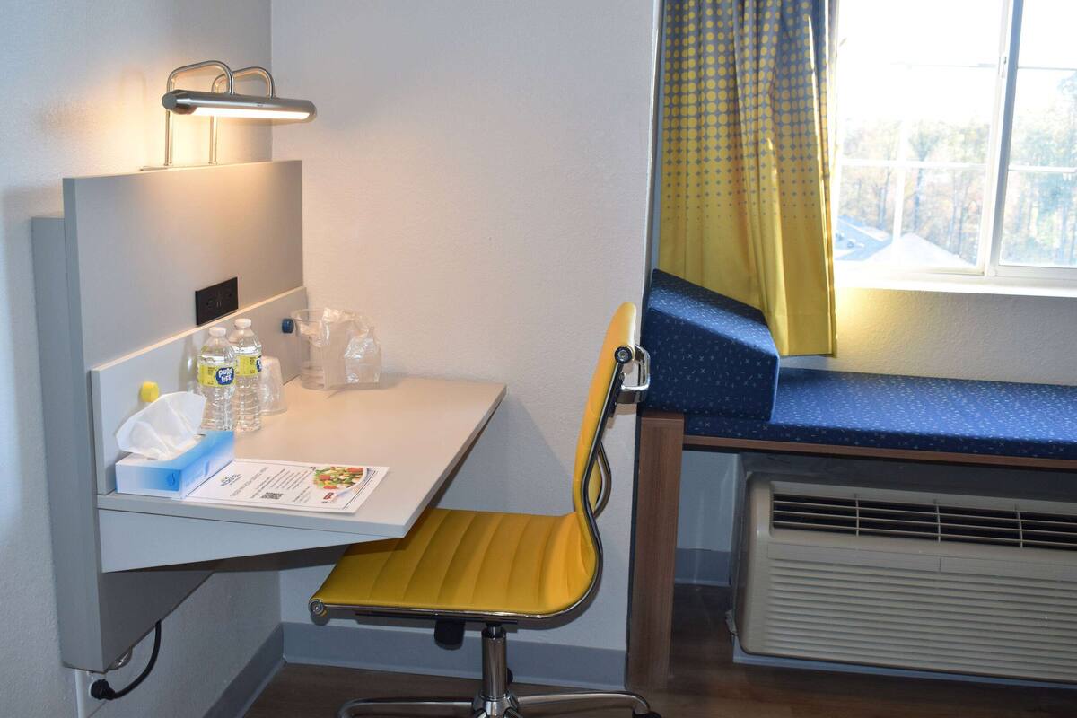 Une petite chambre d’hôtel avec un bureau, une chaise et une fenêtre avec des rideaux.