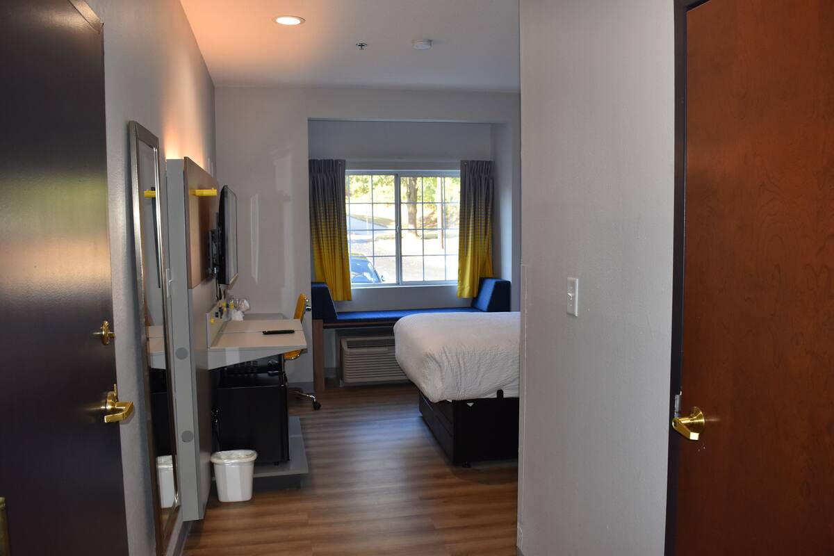 Une chambre d’hôtel avec un lit, un bureau, une chaise, une fenêtre avec des rideaux et une porte.