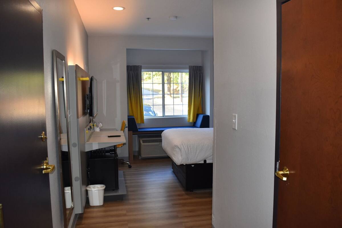 Une chambre d’hôtel avec un lit, un bureau et une chaise, une fenêtre avec des rideaux et une porte.