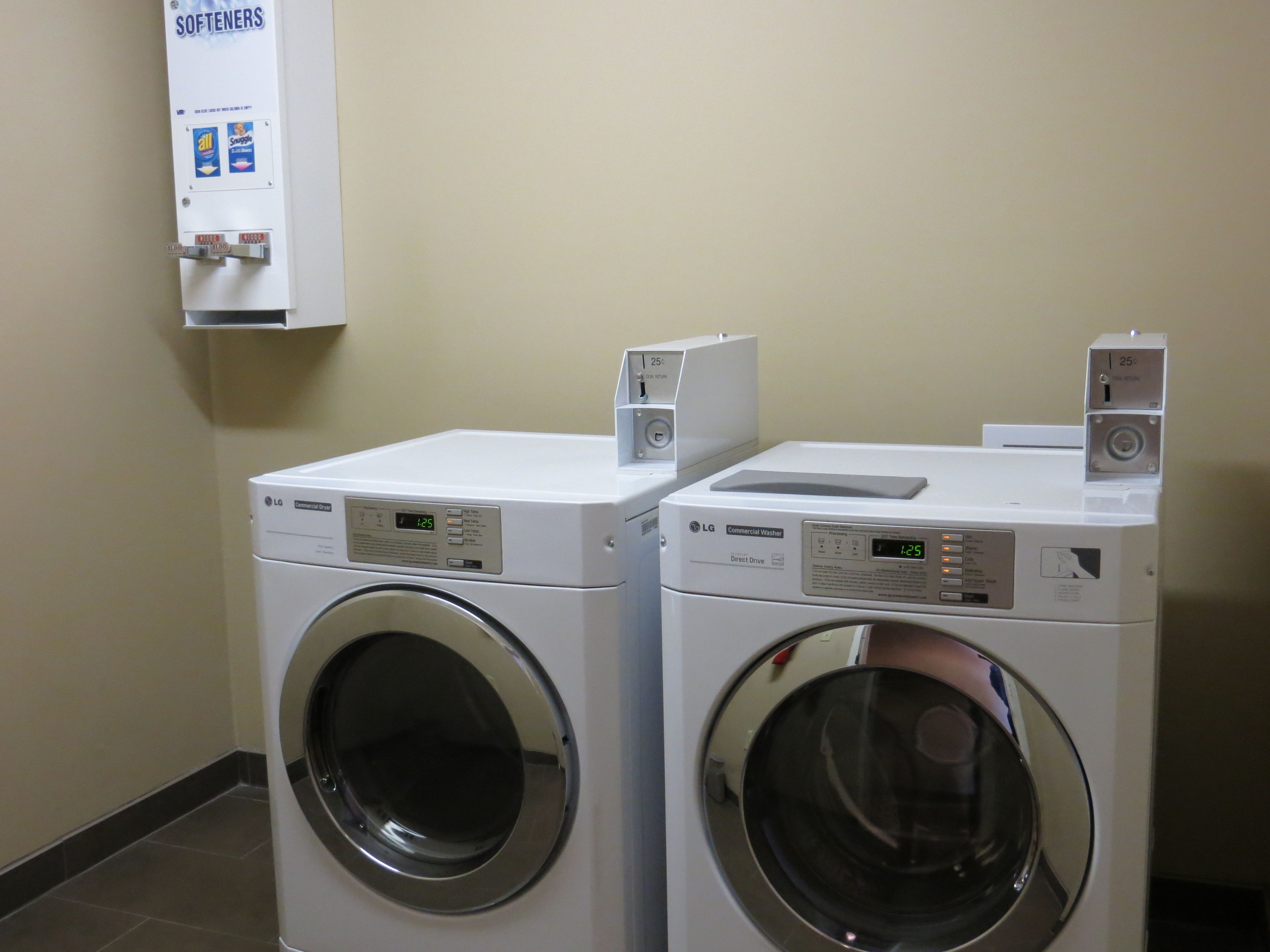 Two white LG commercial washers with digital displays, a wall-mounted softener dispenser, and a tiled floor.