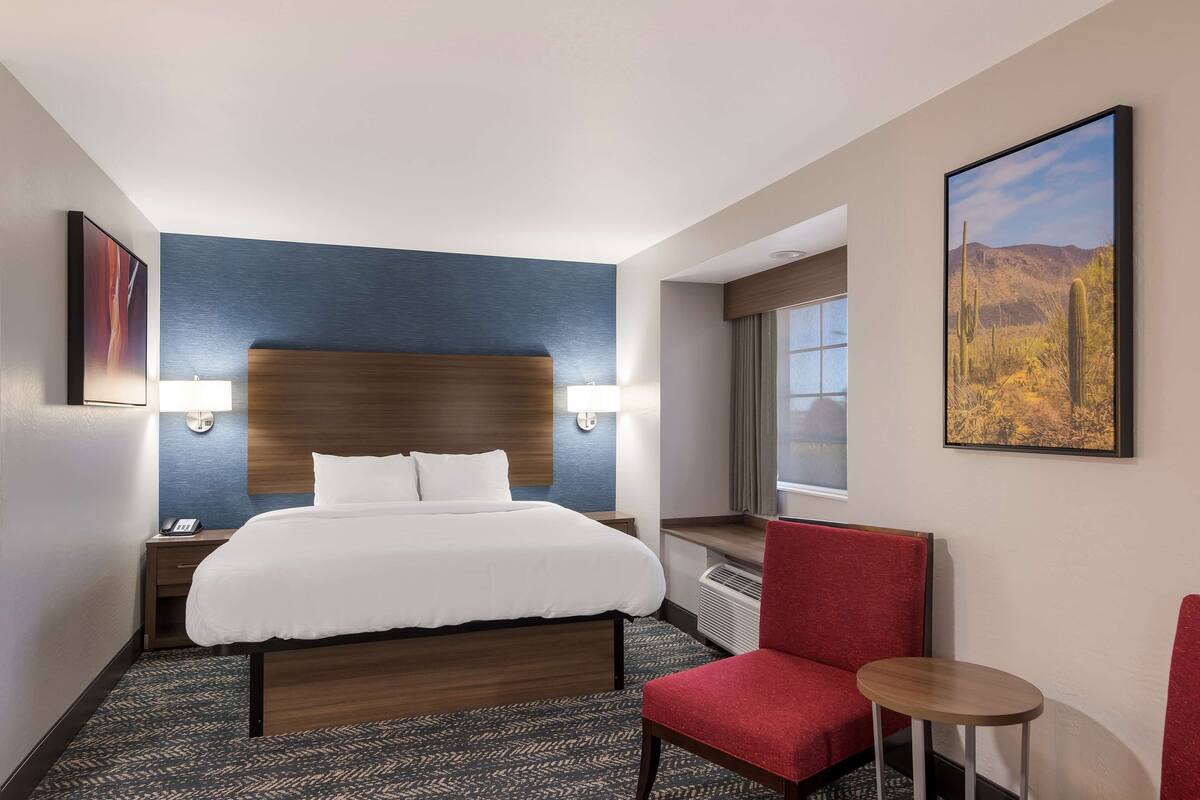 A hotel room with a large bed, a red chair, a small table, and a window with a view of a desert landscape.