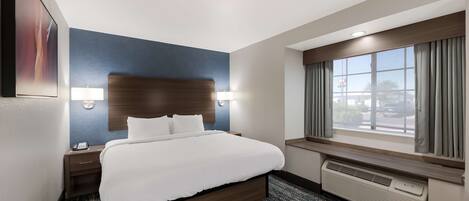 A hotel room with a large bed, a nightstand, a wall-mounted lamp, a window with curtains, and a wall-mounted air conditioner.