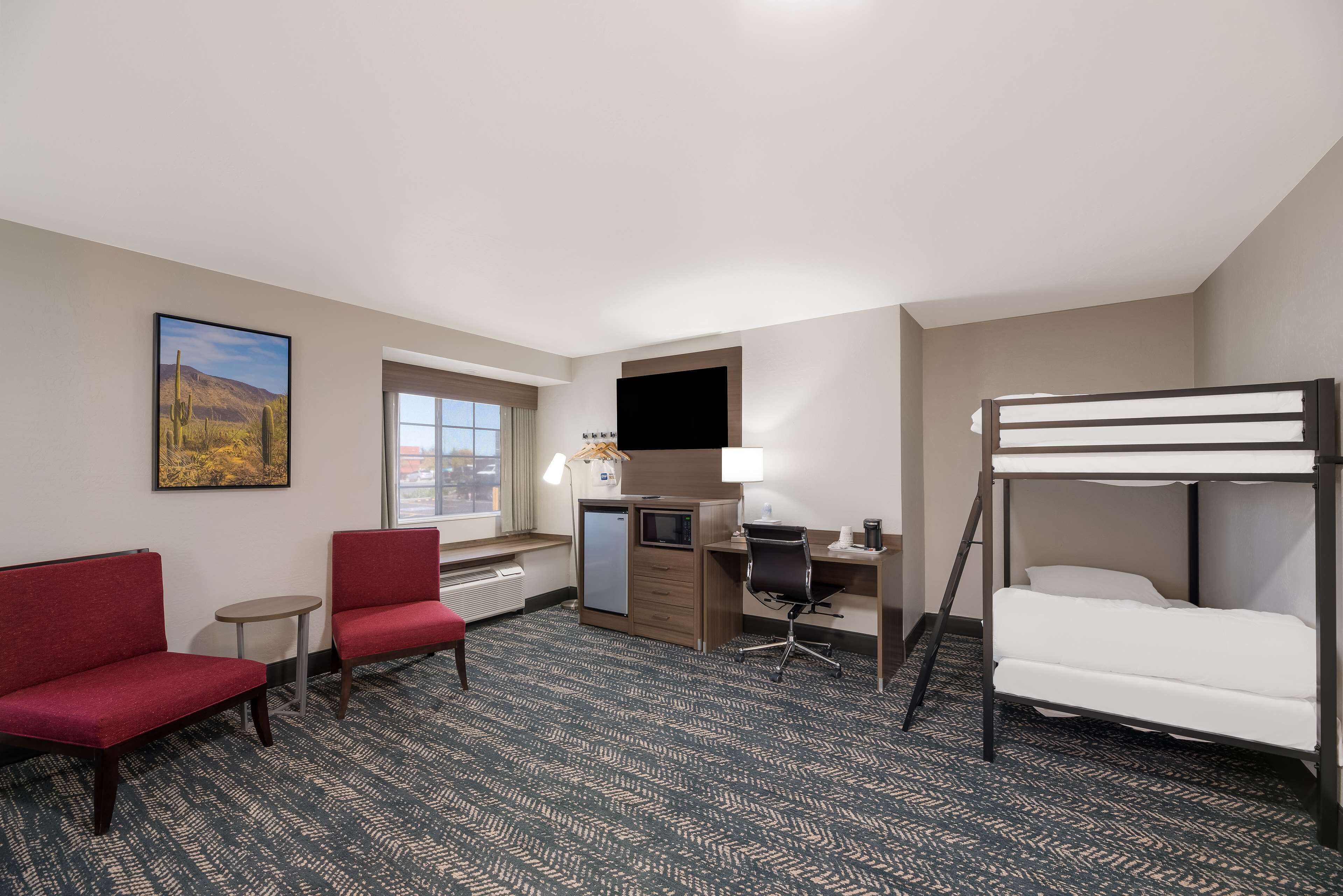 A hotel room with a bunk bed, a desk with a computer, two red chairs, a small table, and a window with a view of the city.