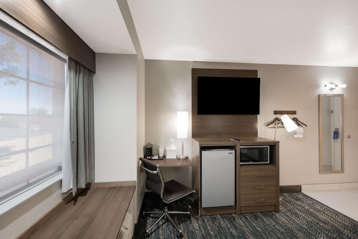 A hotel room with a desk, chair, flat-screen TV, mini-fridge, and a large window with curtains.